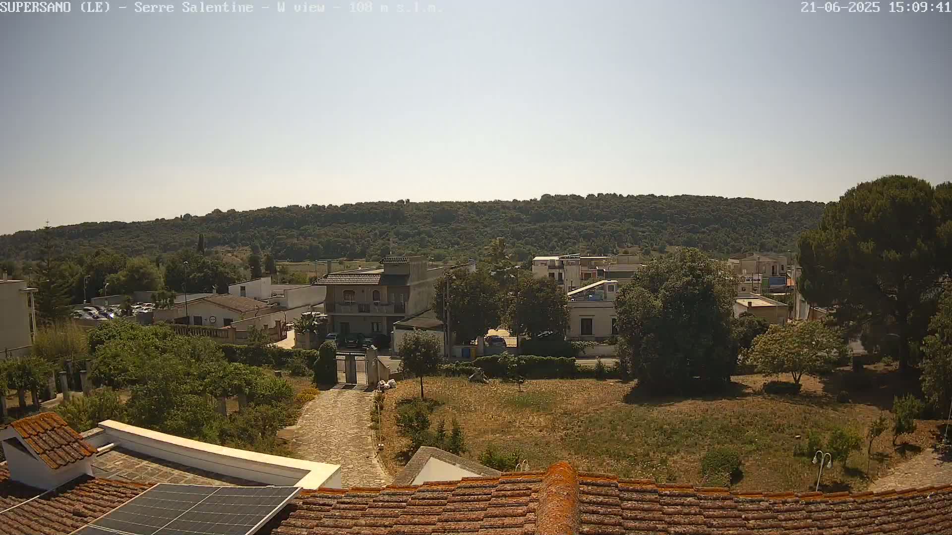 A sunlit view from a rooftop shows a residential area with low buildings and a paved driveway, leading to a gate, set against a backdrop of a forested hill.