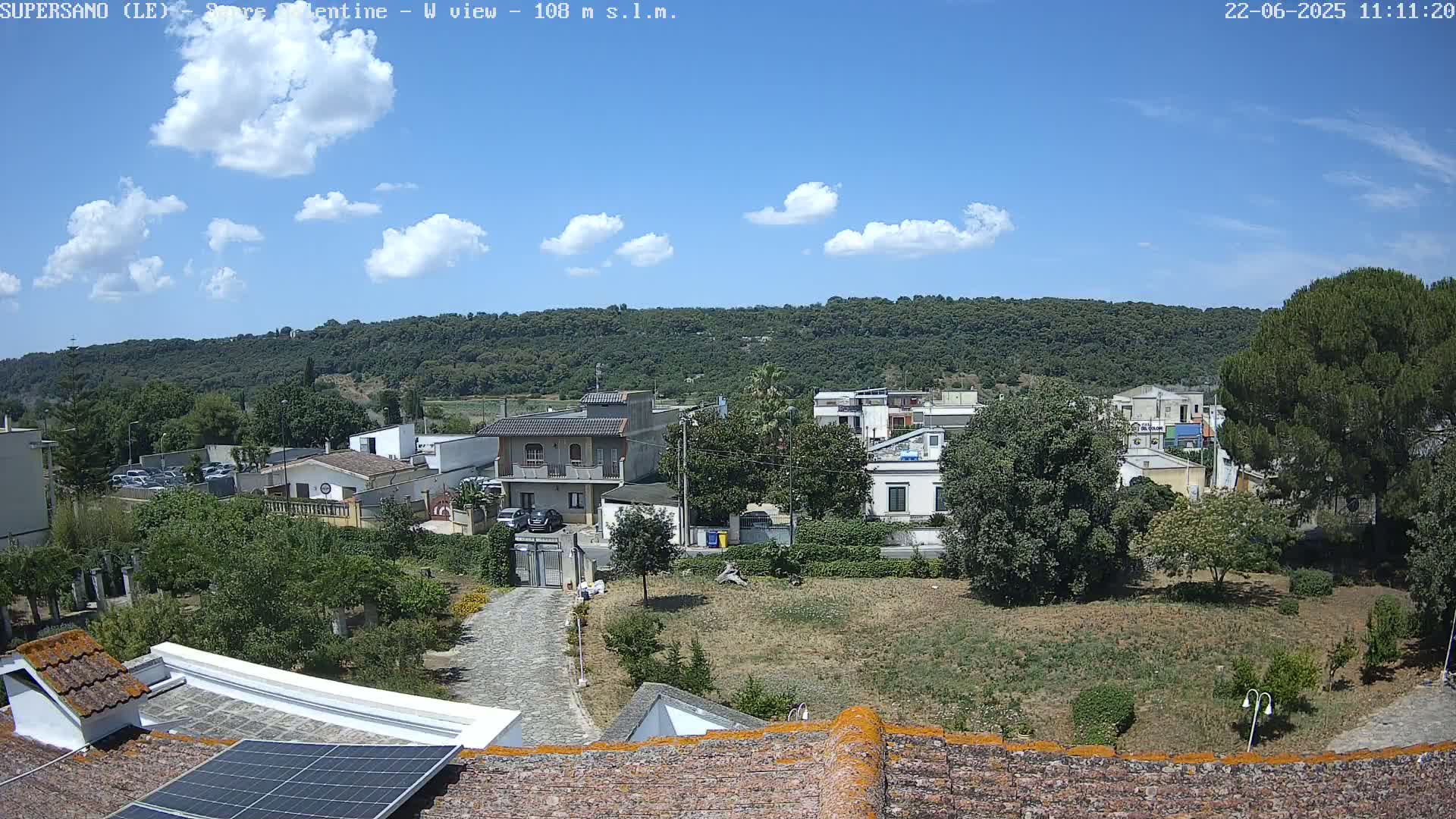 A sunny day reveals a residential area nestled against a verdant hillside, with a few puffy clouds scattered across a clear blue sky.