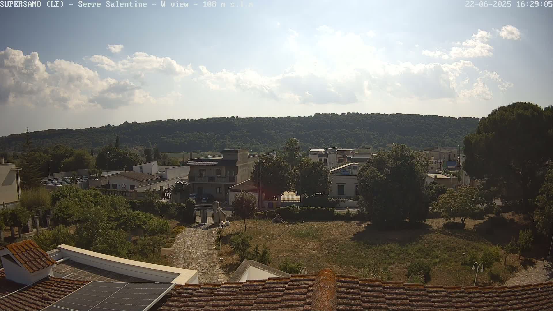 A partly sunny day reveals a residential area nestled beside a verdant hill, with a view from a rooftop showing a stone path leading to buildings and trees.