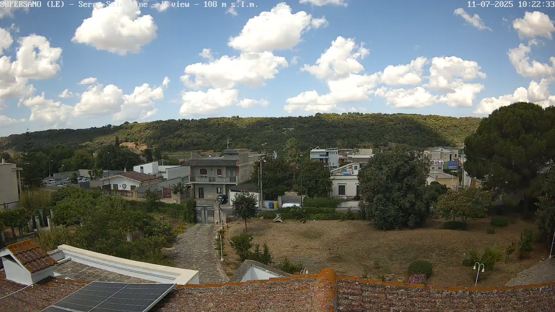 A partly sunny day shows a residential area with low buildings and trees, situated below a hill covered in lush vegetation.
