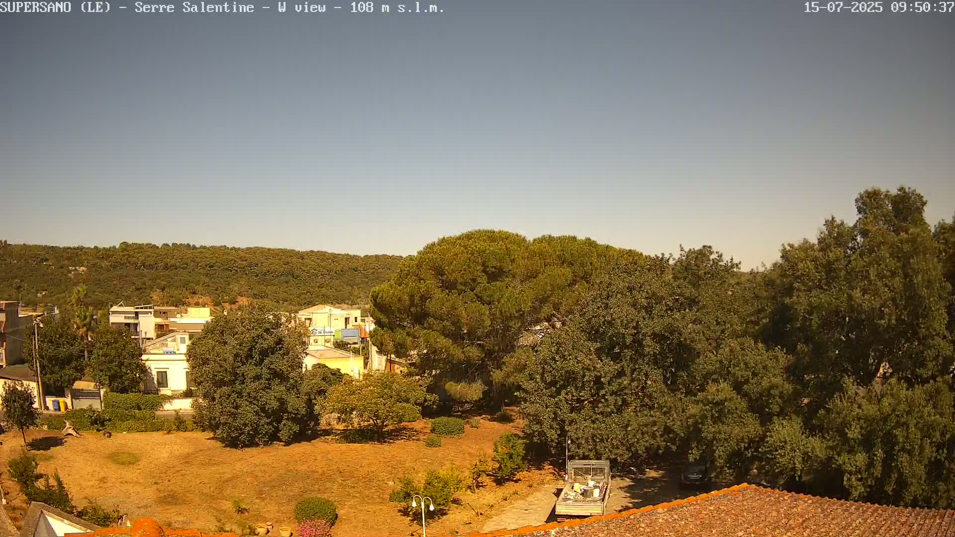 A clear, sunny day reveals a town nestled amongst green hills and lush vegetation, with a few buildings and vehicles visible from a high vantage point.