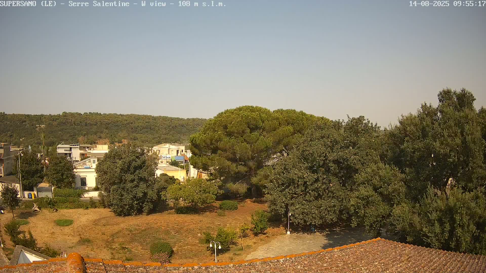 A mostly clear, sunny day reveals a view of a town nestled amongst green, leafy trees and hills.