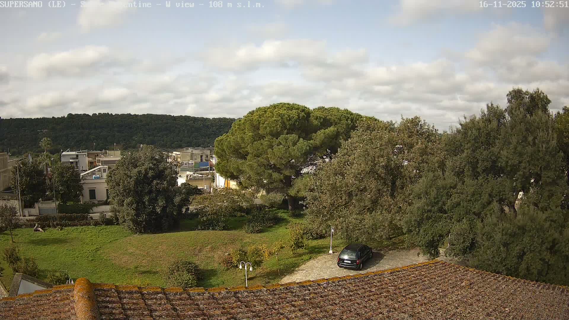 A partly cloudy sky hangs over a tree-covered hillside and a town with various buildings and lush greenery, including a grassy area and a parked car, viewed from above a tiled roof.