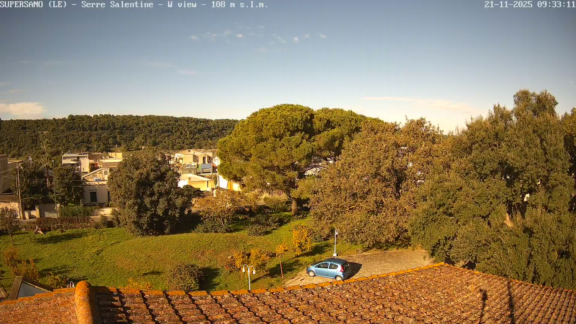 A clear blue sky with a few clouds presides over an elevated view of a rural landscape, showcasing a tiled rooftop in the foreground, a verdant hillside with a blue car and diverse trees, a cluster of houses, and a dense forested hill in the background.