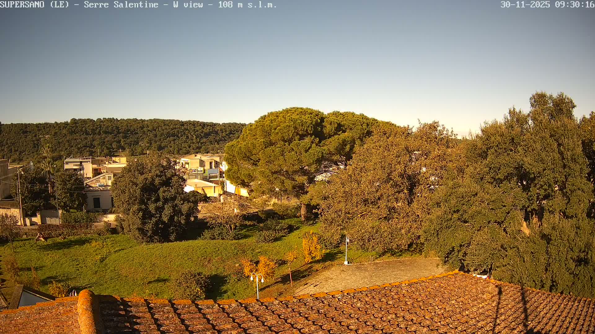 An outdoor scene on a clear and sunny day features a terracotta tiled roof in the foreground, looking over a green hillside dotted with houses and trees, towards a dense forest under a bright blue sky.