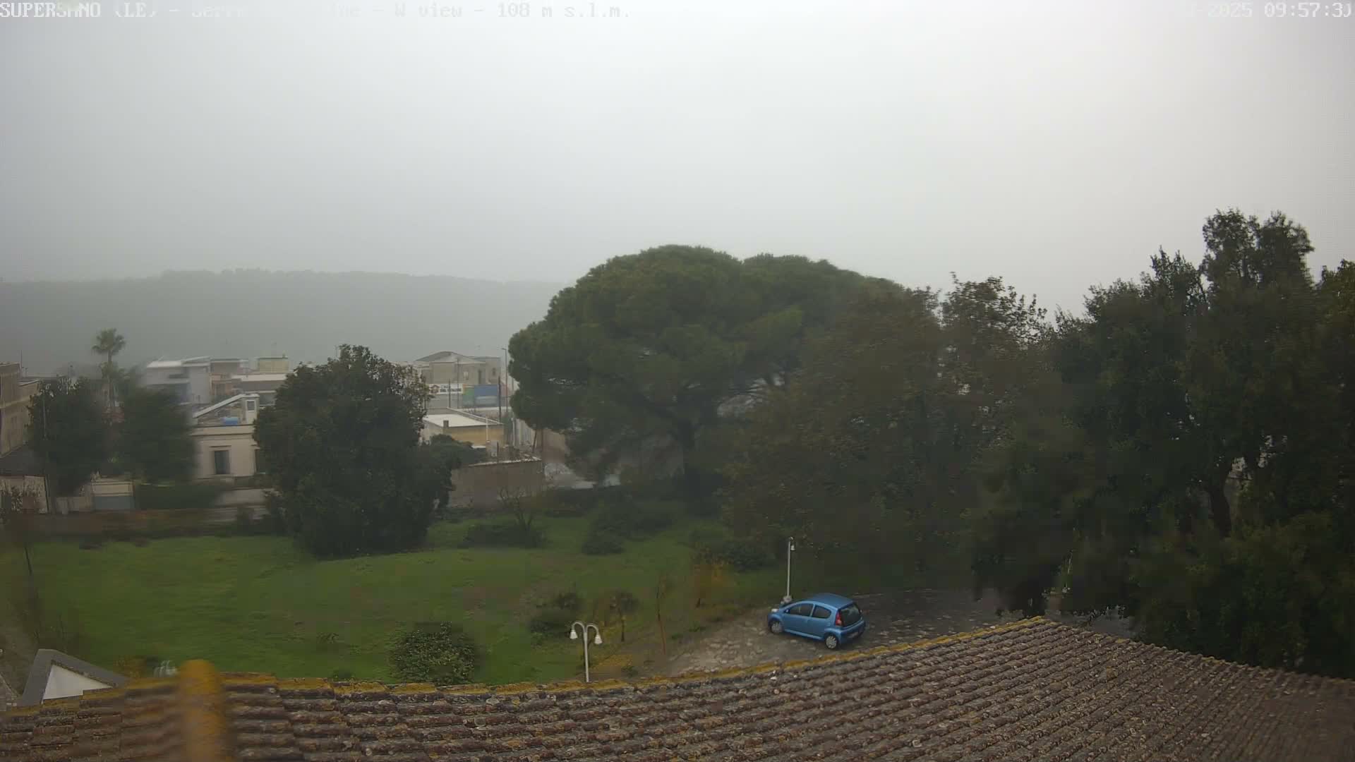 A panoramic view from above a mossy tiled roof reveals a misty, overcast day over a town with green spaces, scattered trees, a parked blue car, and hazy hills in the distance.