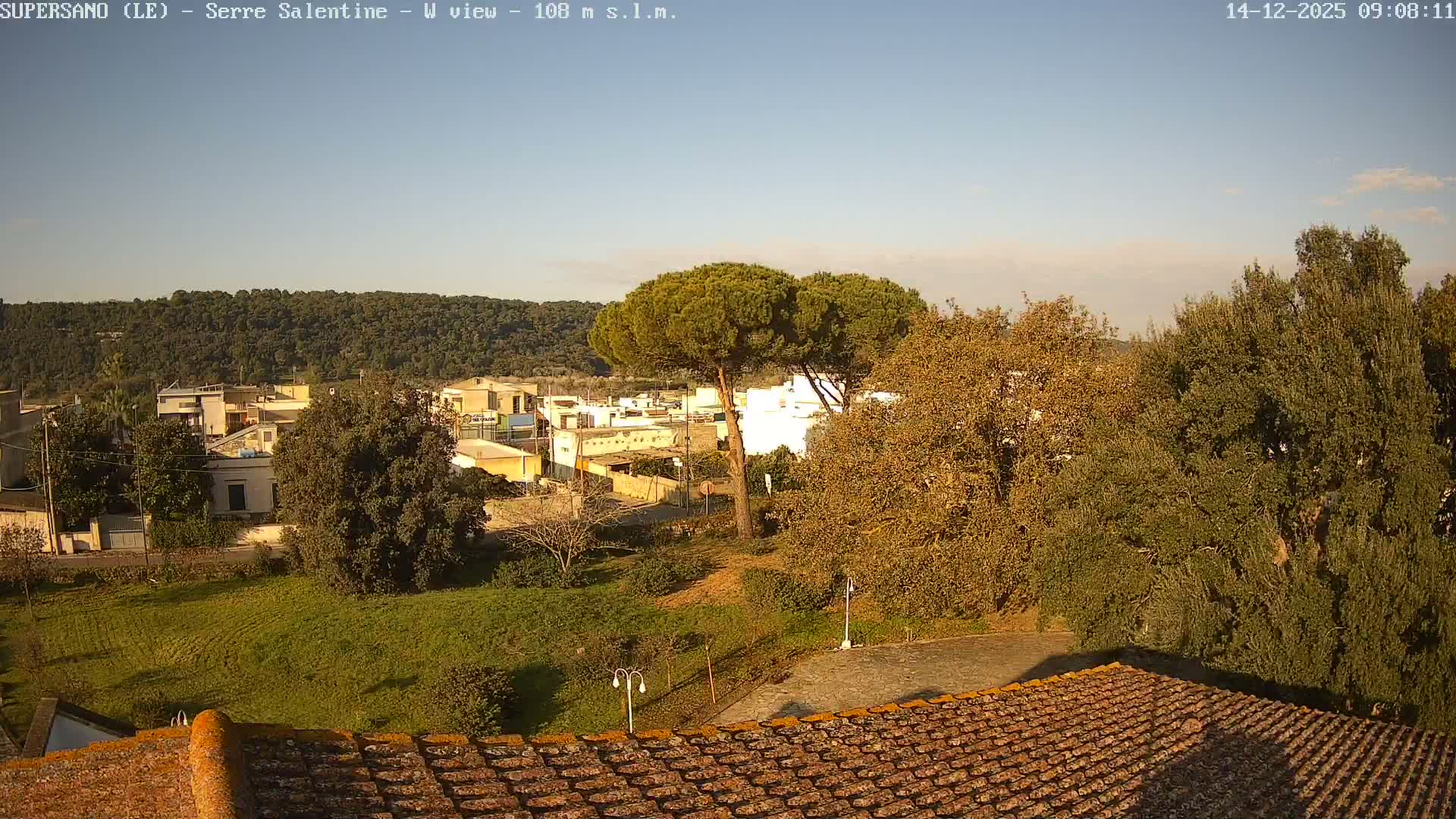 On a clear and sunny day, the scene overlooks a terracotta tiled roof towards a verdant landscape with various trees, a village, and a densely forested hill in the background.