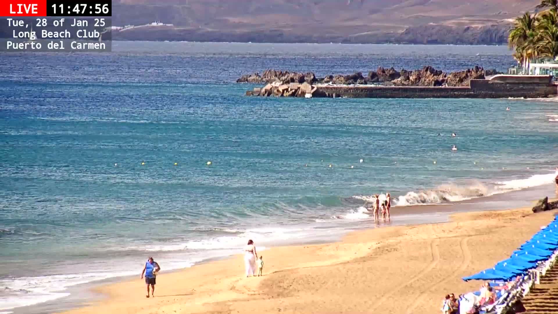 A sandy beach with a few people walking and some in the water, under a sunny sky, next to a calm ocean with some small waves.