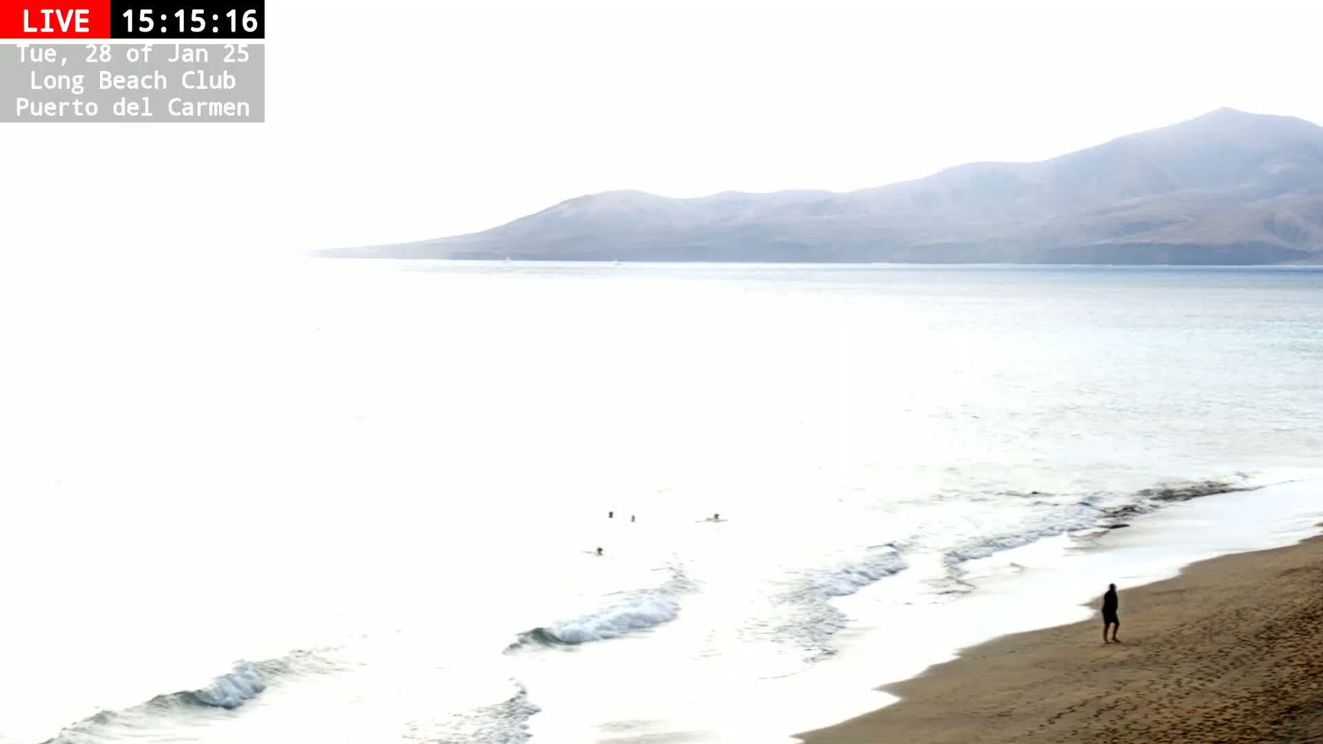 A person stands on a sandy beach next to gentle waves lapping the shore, with a hazy mountain range visible in the distance under a bright, mostly clear sky.