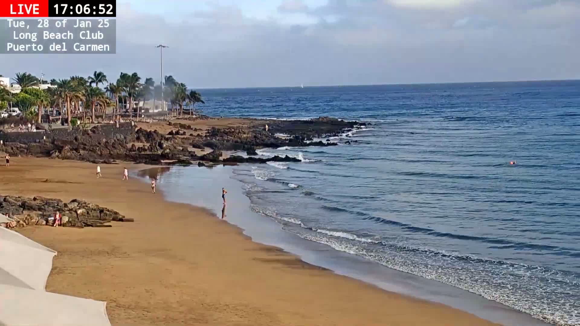 A sandy beach with a few people on it meets a calm, blue ocean with small waves under a partly cloudy sky.