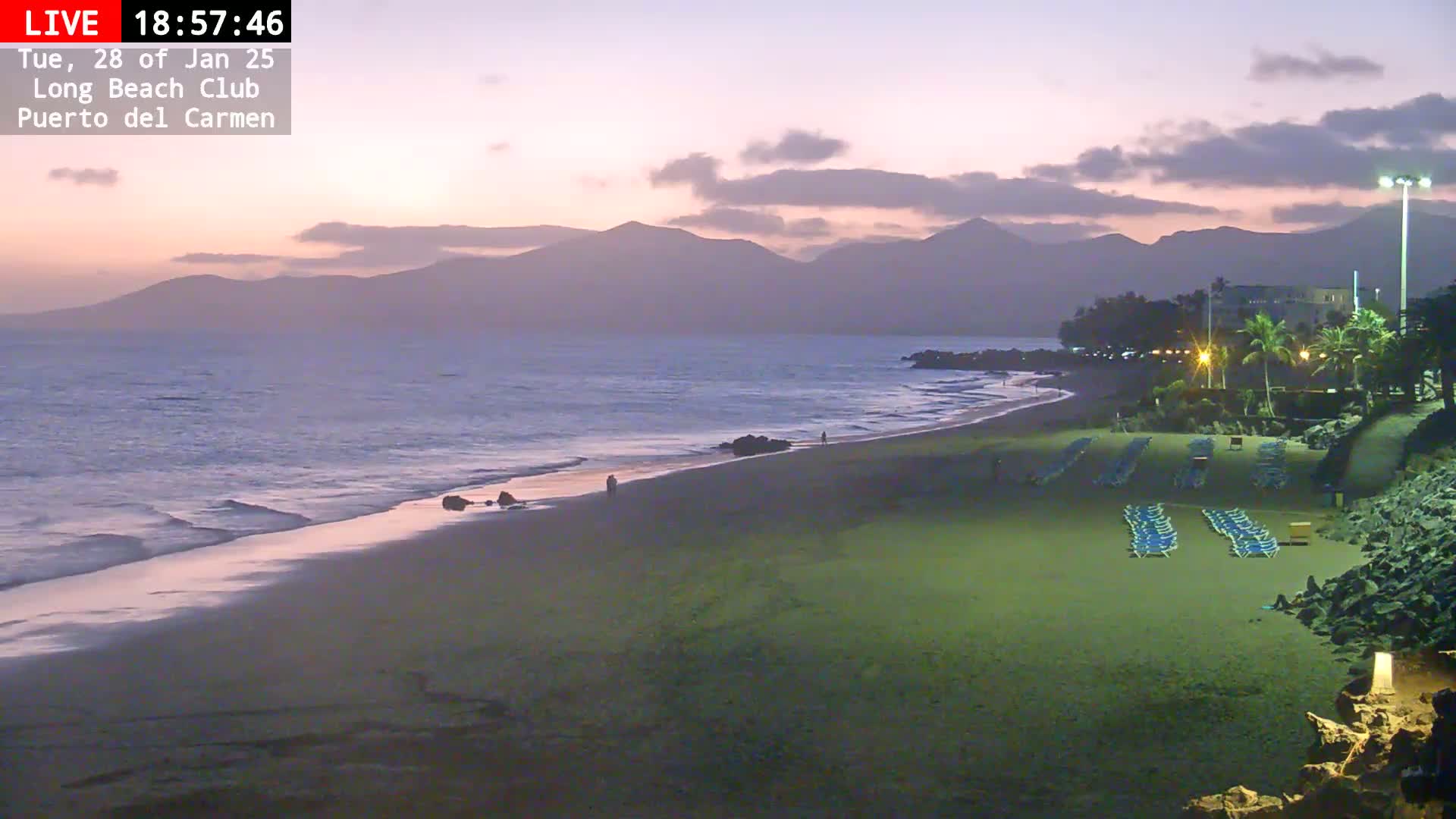 A sandy beach with rows of lounge chairs sits under a twilight sky, with calm waves lapping the shore and mountains visible in the distance.