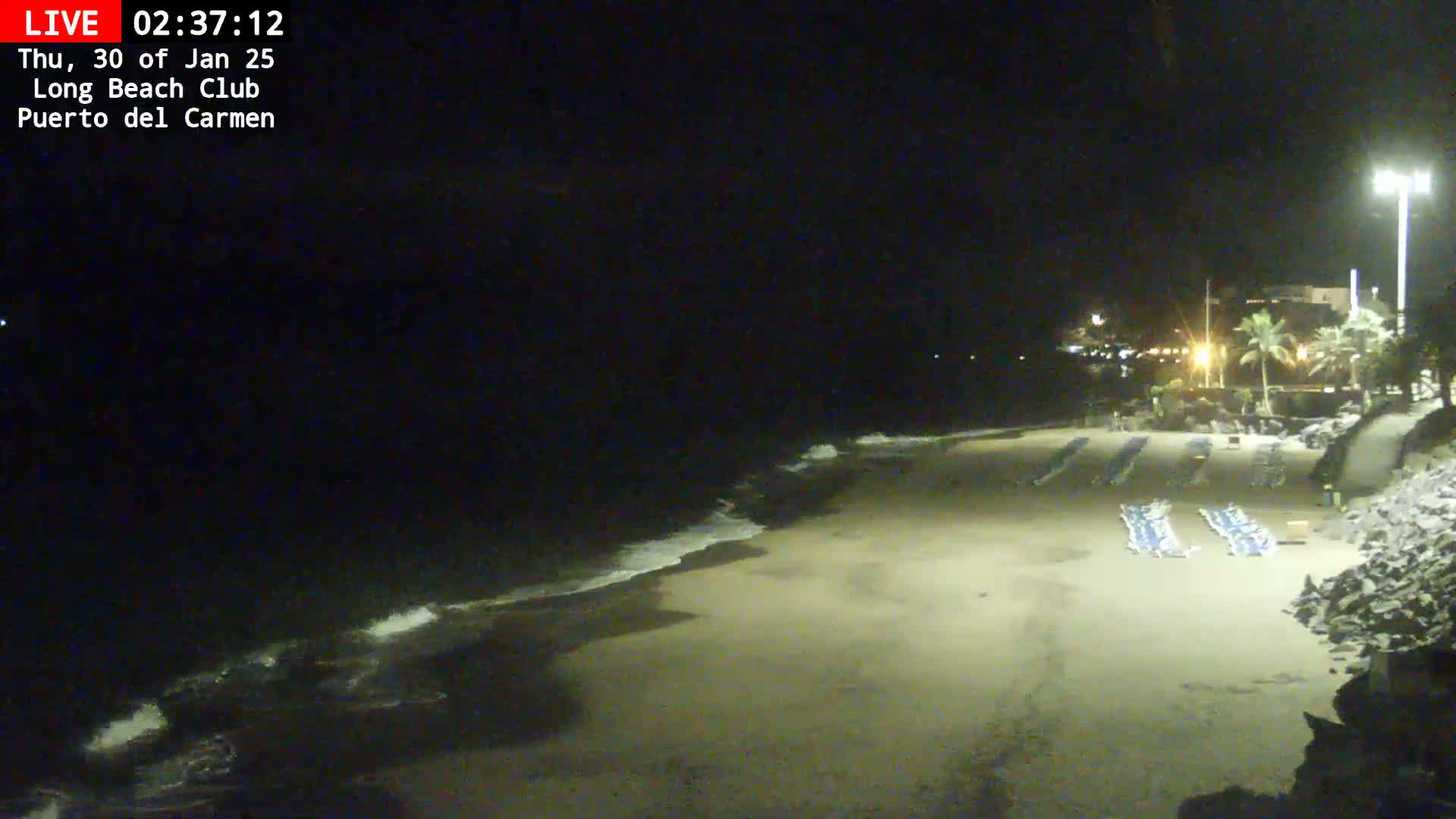 A dark, sandy beach at night with gentle waves lapping the shore, a few folded lounge chairs visible on the sand, and a dimly lit area beyond.