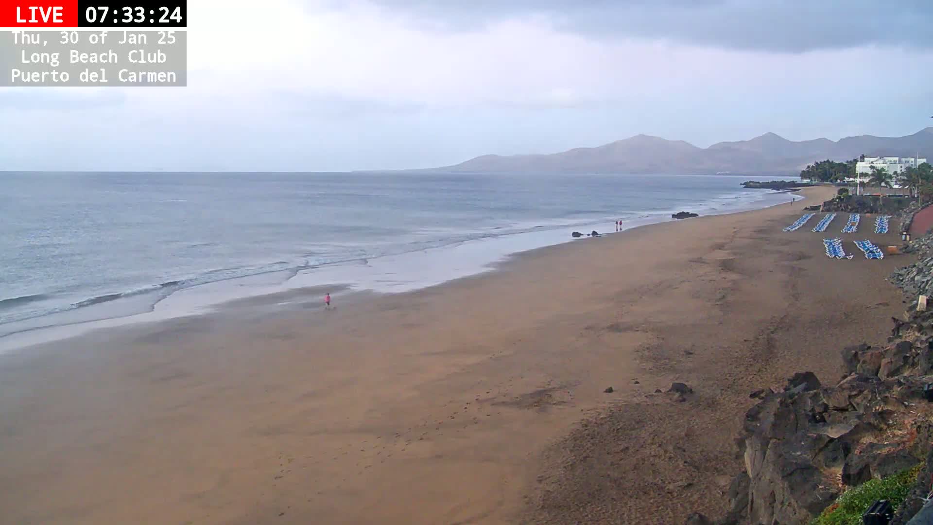 A mostly empty sandy beach stretches along a calm ocean under an overcast sky, with a few people visible and rows of beach chairs lined up near some buildings in the distance.
