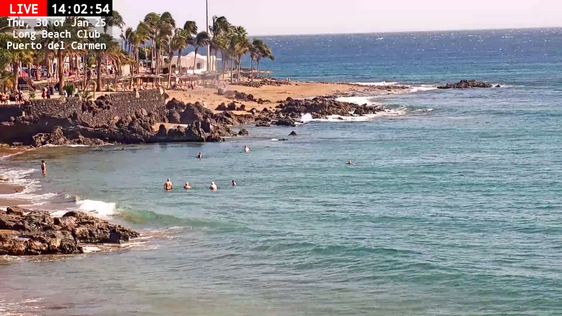 A partly sunny day at a rocky beach with calm, teal-colored water where several people are swimming near the shore, with palm trees visible on the sand in the background.