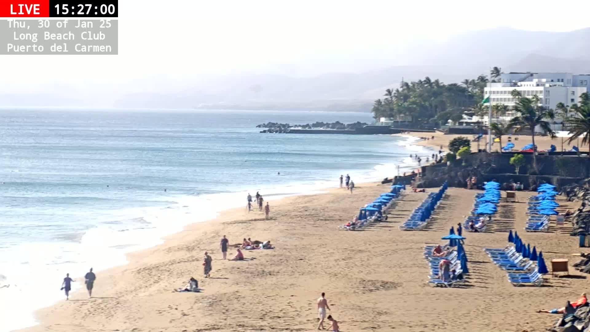 A hazy beach scene shows a sandy shore with scattered people, rows of blue beach umbrellas and lounge chairs, gentle waves lapping the sand, and a hotel in the background.