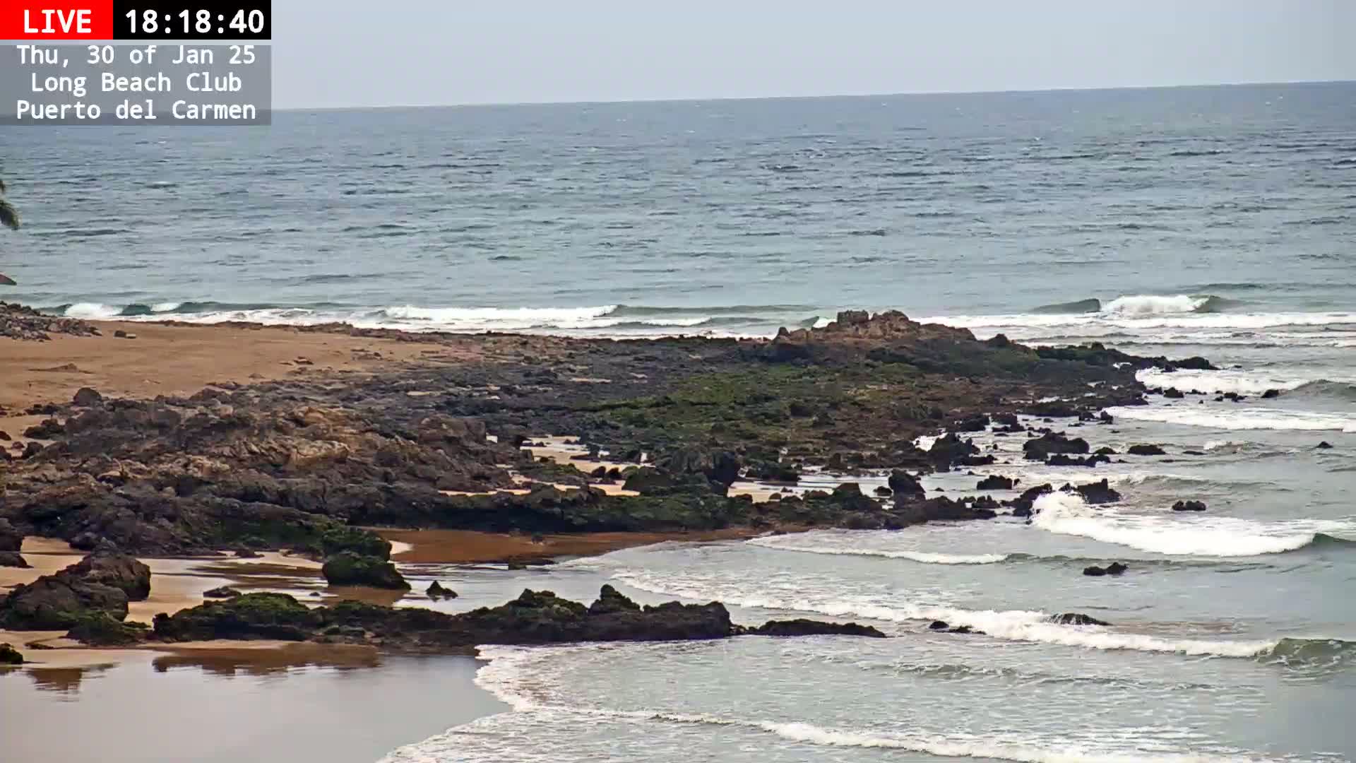 A rocky shoreline with a sandy beach meets a calm ocean under a mostly clear sky.