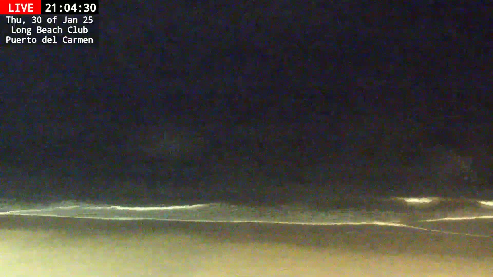 A dark night scene shows a beach with gently lapping waves under a mostly clear, dark sky.