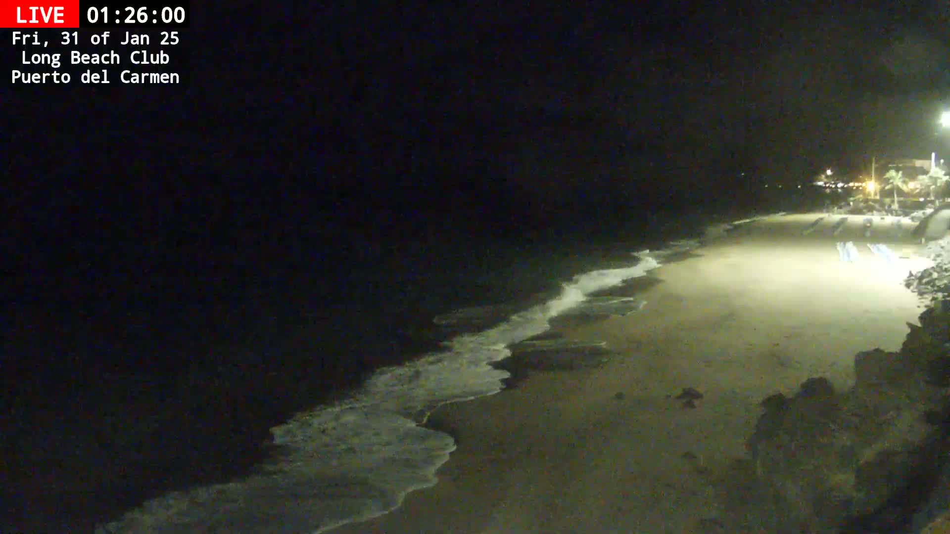 A mostly empty sandy beach at night is gently lapped by small waves under a dark sky.
