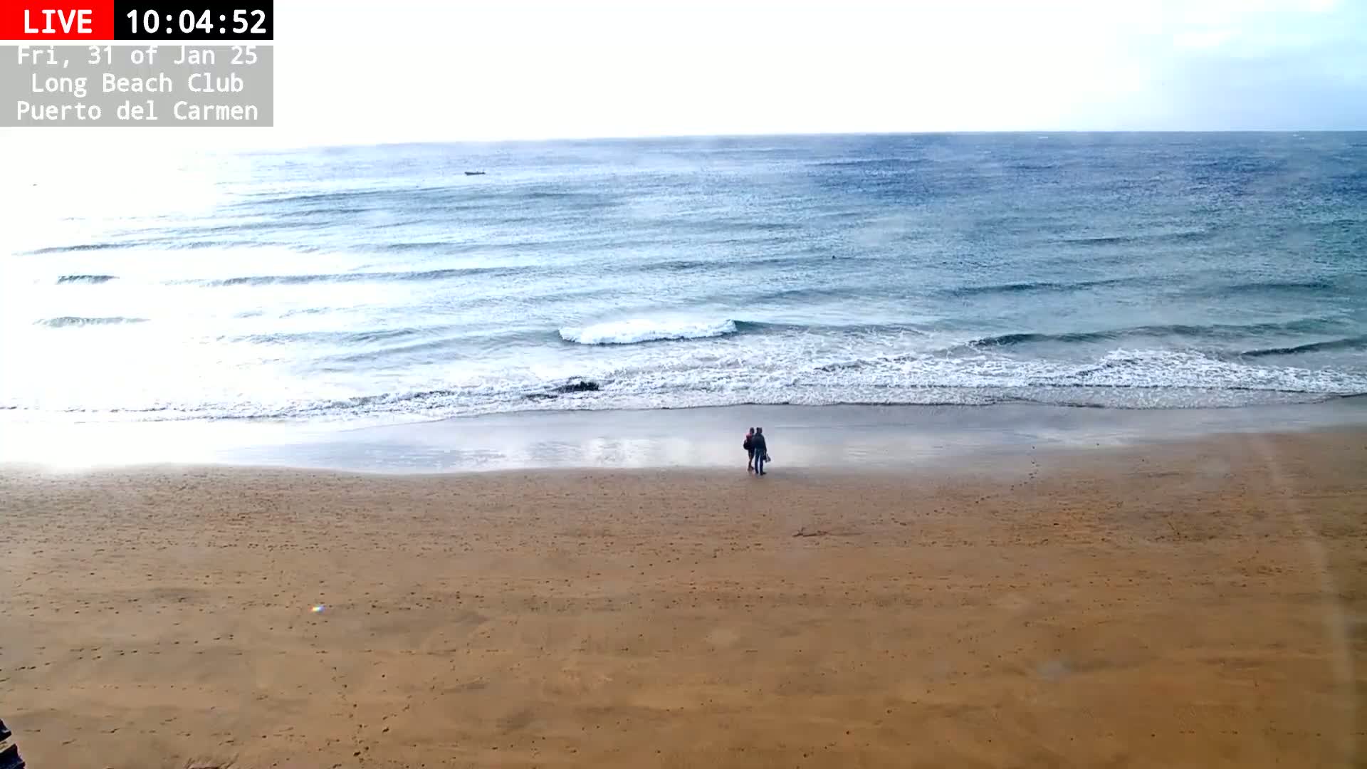 A sandy beach with a couple walking along the shoreline, gentle waves lapping the shore under an overcast sky.