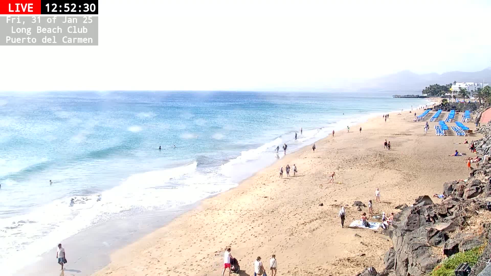 A sandy beach with scattered people, blue lounge chairs, and gentle waves under a hazy sky.