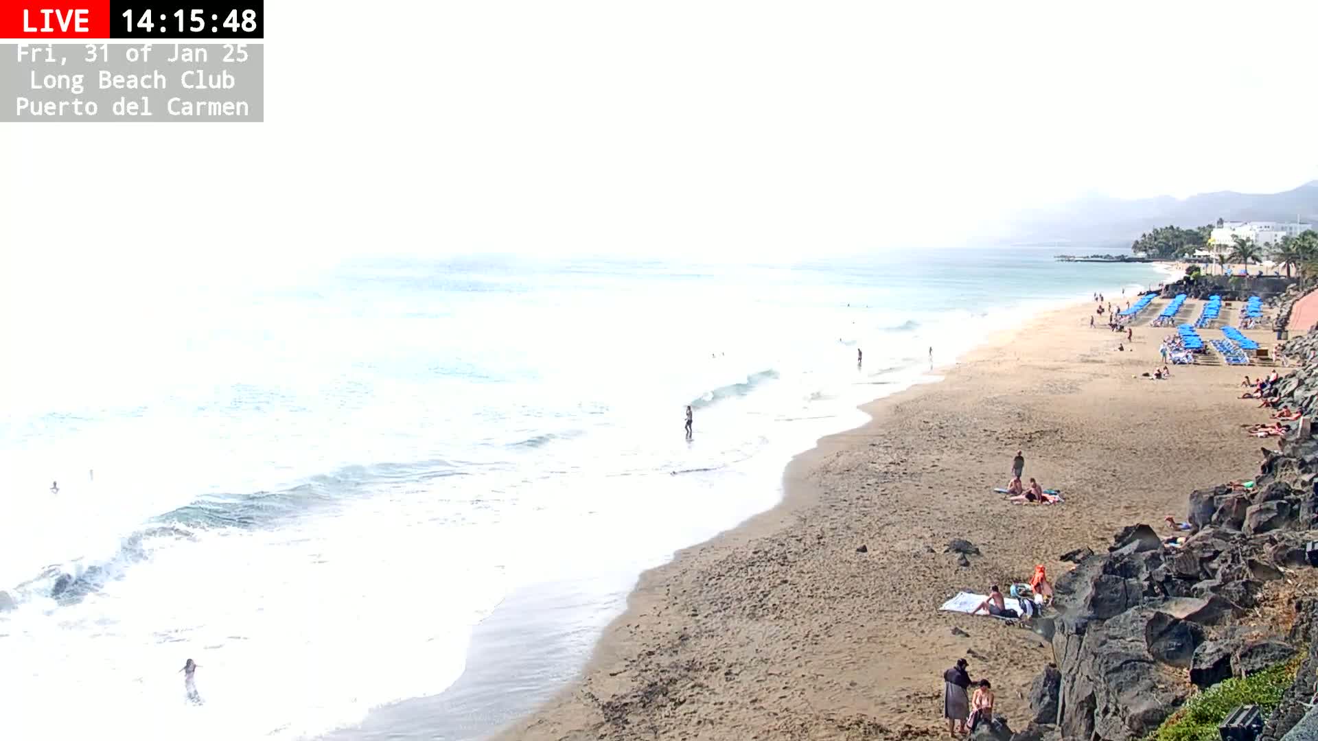A sandy beach with a few people sunbathing and swimming in calm, light-blue ocean water under an overcast sky.