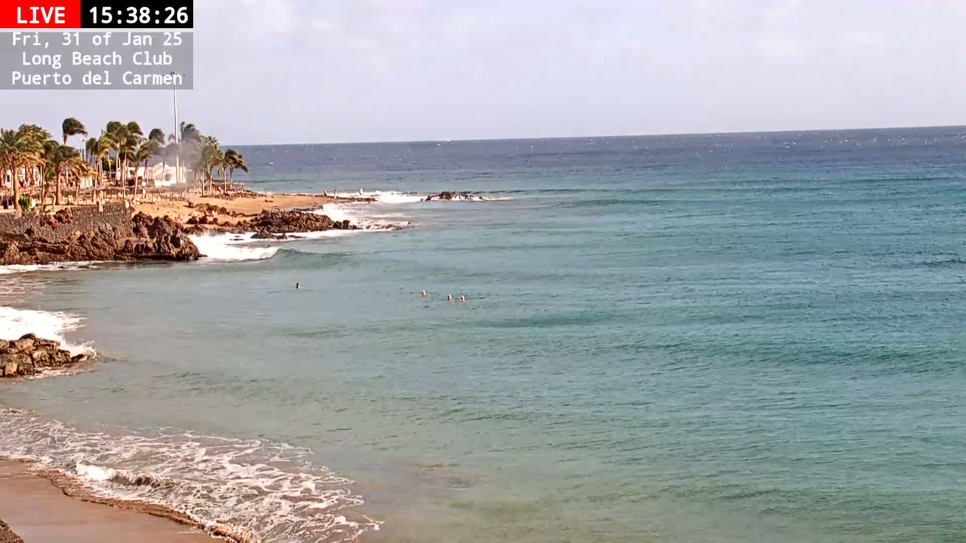A sandy beach with calm, clear turquoise water, gentle waves, and a few people swimming, under a mostly sunny sky.