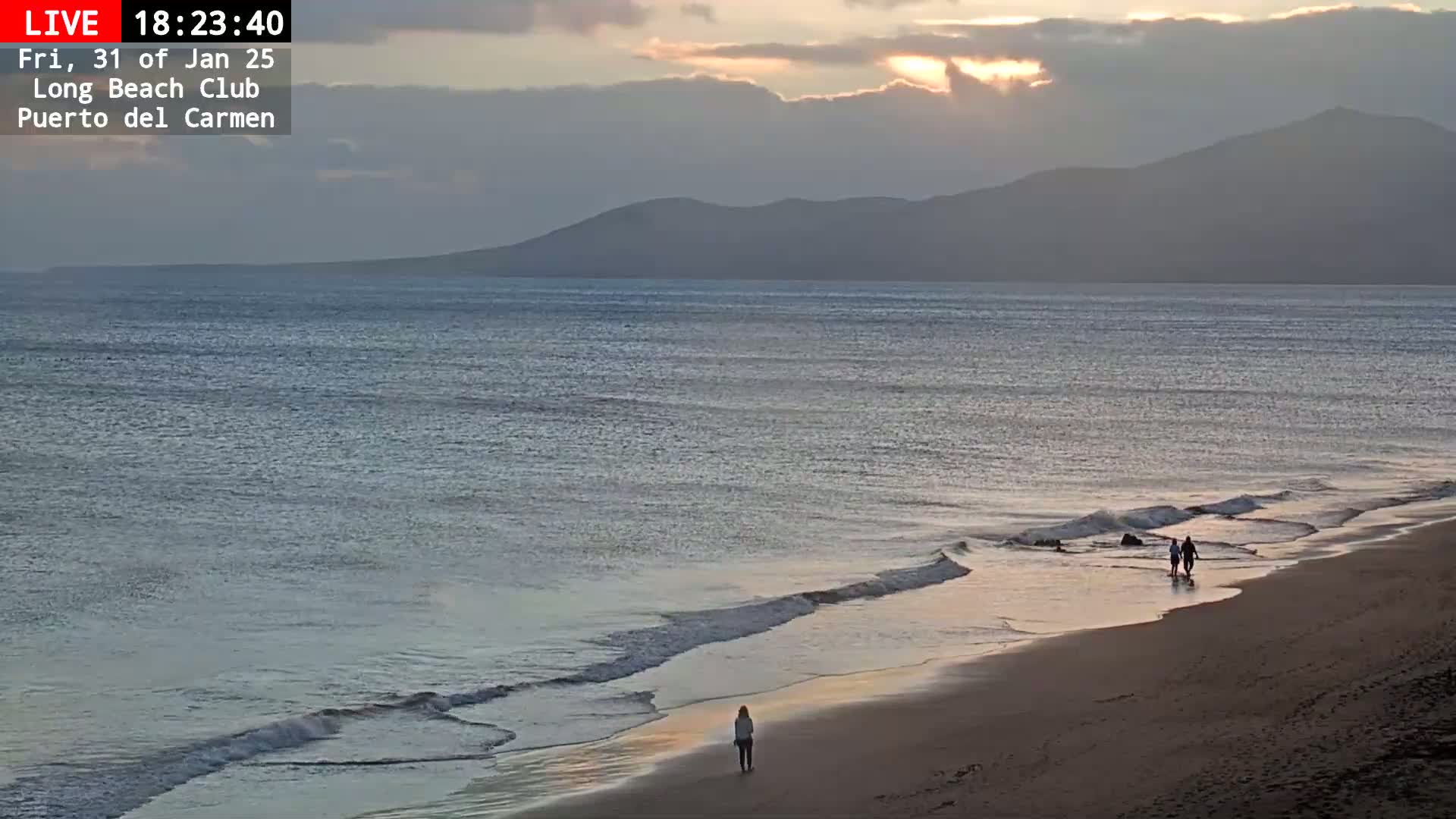 A woman walks alone on a sandy beach as the sun sets over calm ocean waters and distant mountains under a cloudy sky.