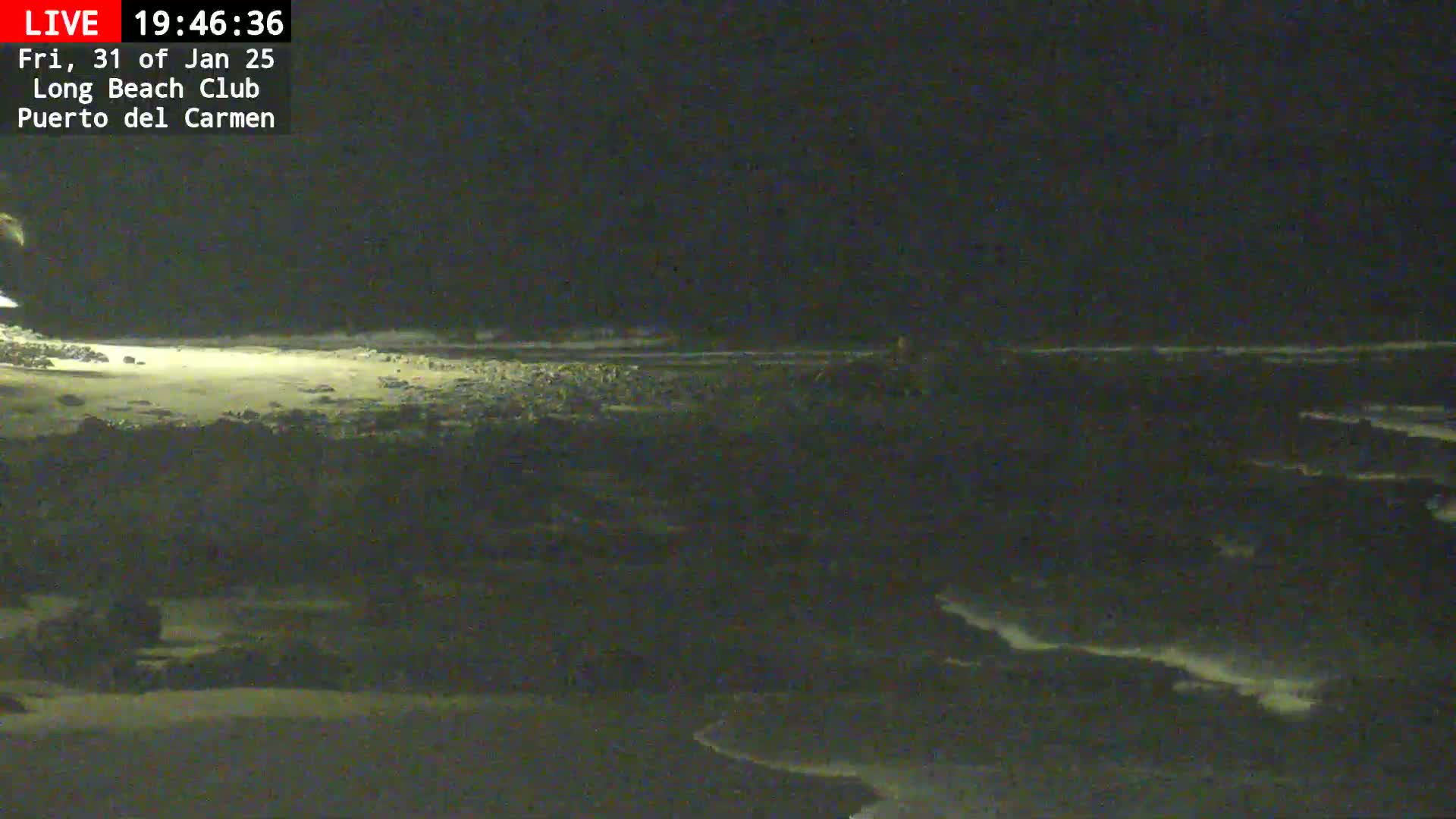 A dark, nighttime beach scene shows a sandy shore, rocky shoreline, and gently rolling ocean waves under a clear, starless sky.