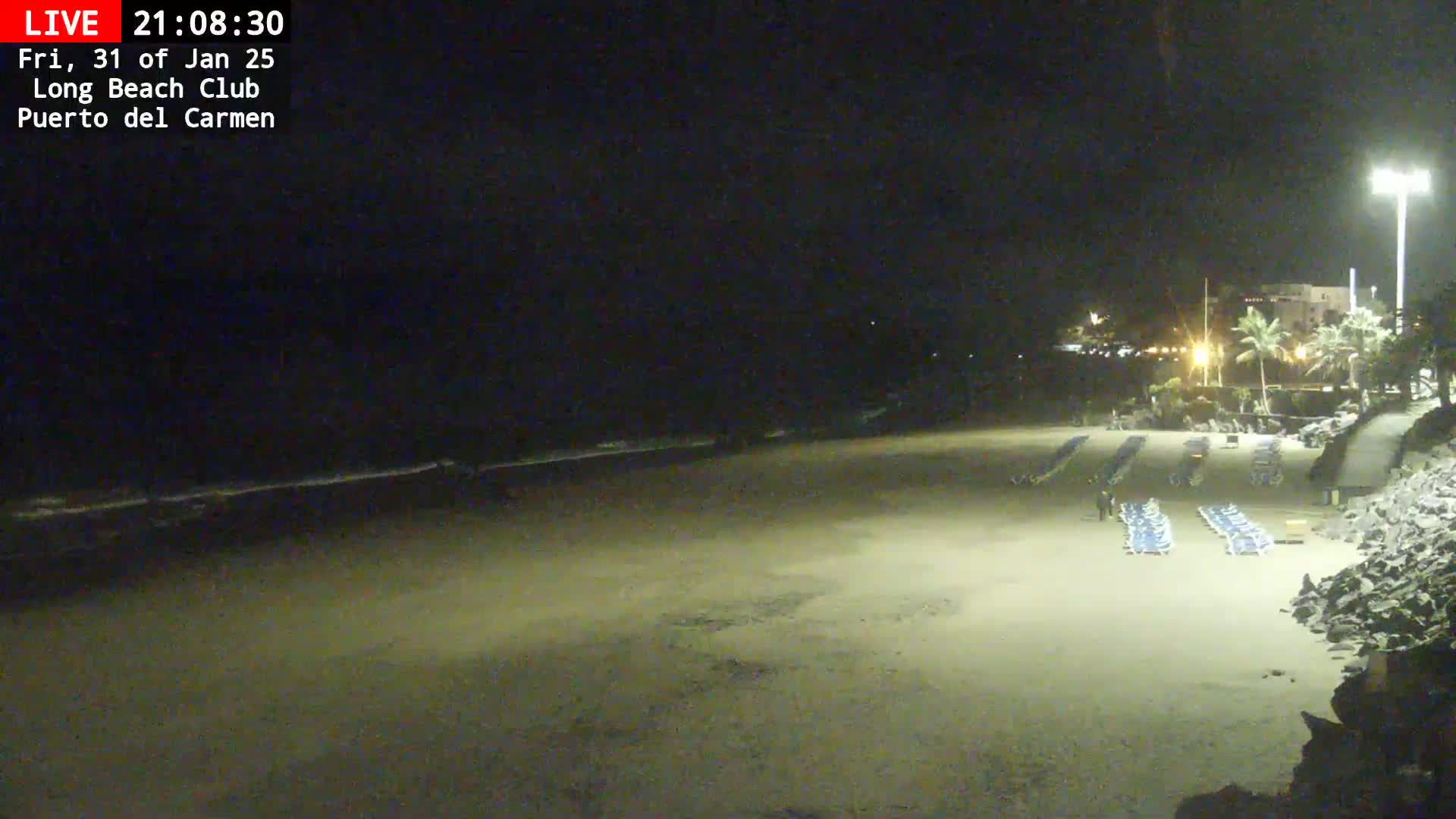 A mostly empty sandy beach at night is lit by artificial light, with a few lounge chairs and a person visible, and a dark ocean in the background.