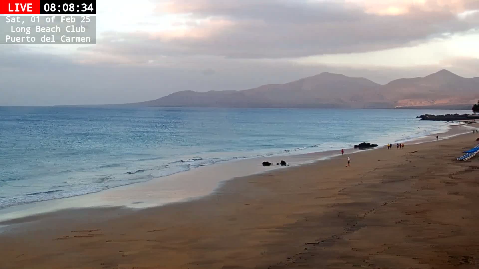 A mostly cloudy sunset illuminates a sandy beach with several people walking along the shoreline and mountains in the distance.