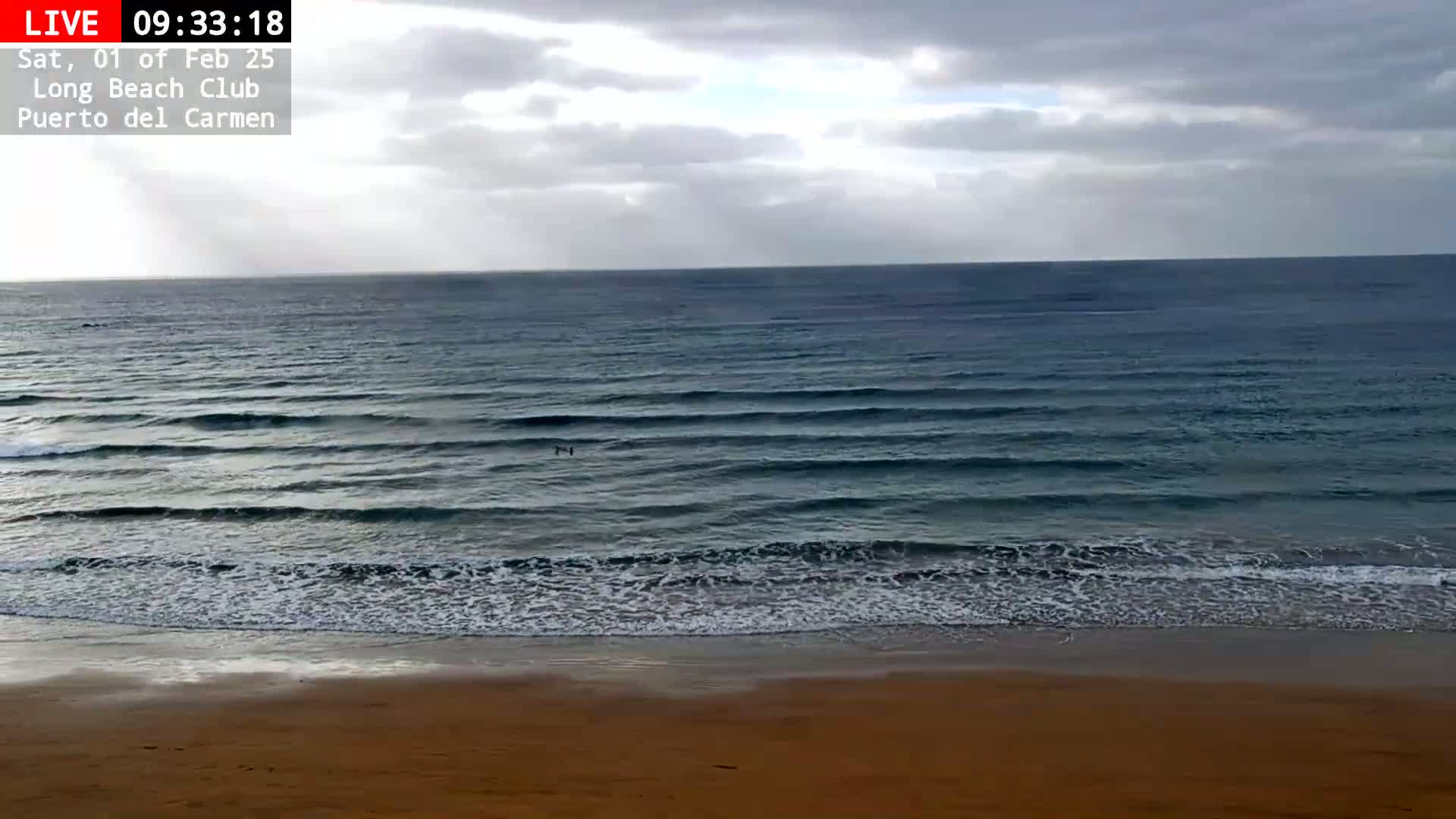 A sandy beach meets a dark-blue ocean with small waves under a mostly cloudy sky.