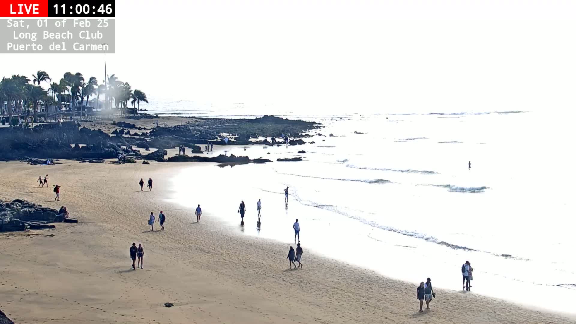 A sandy beach with scattered people walking along the shore, under a bright, hazy sky, features calm waves gently lapping the beach.