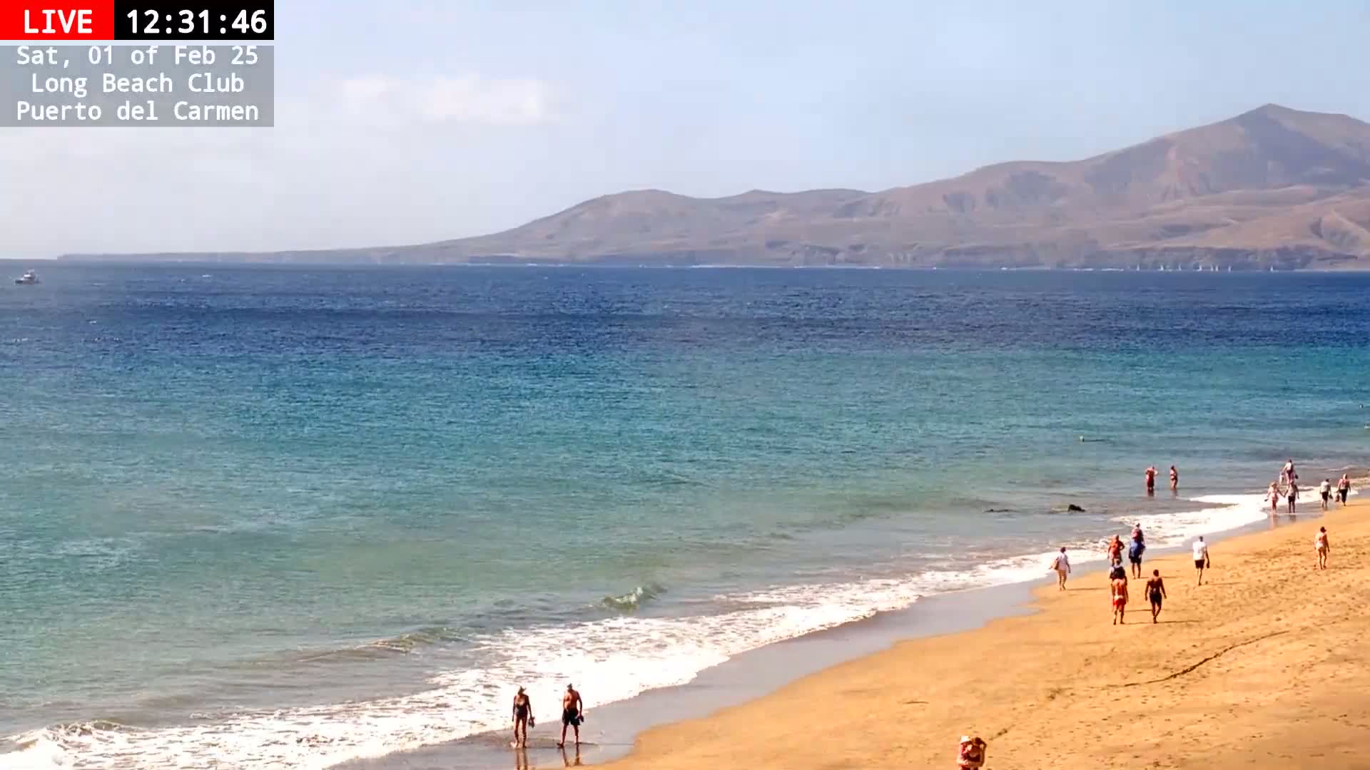 A sandy beach with several people walking along the shore and in the water, under a sunny sky, with a calm ocean and distant mountains visible in the background.