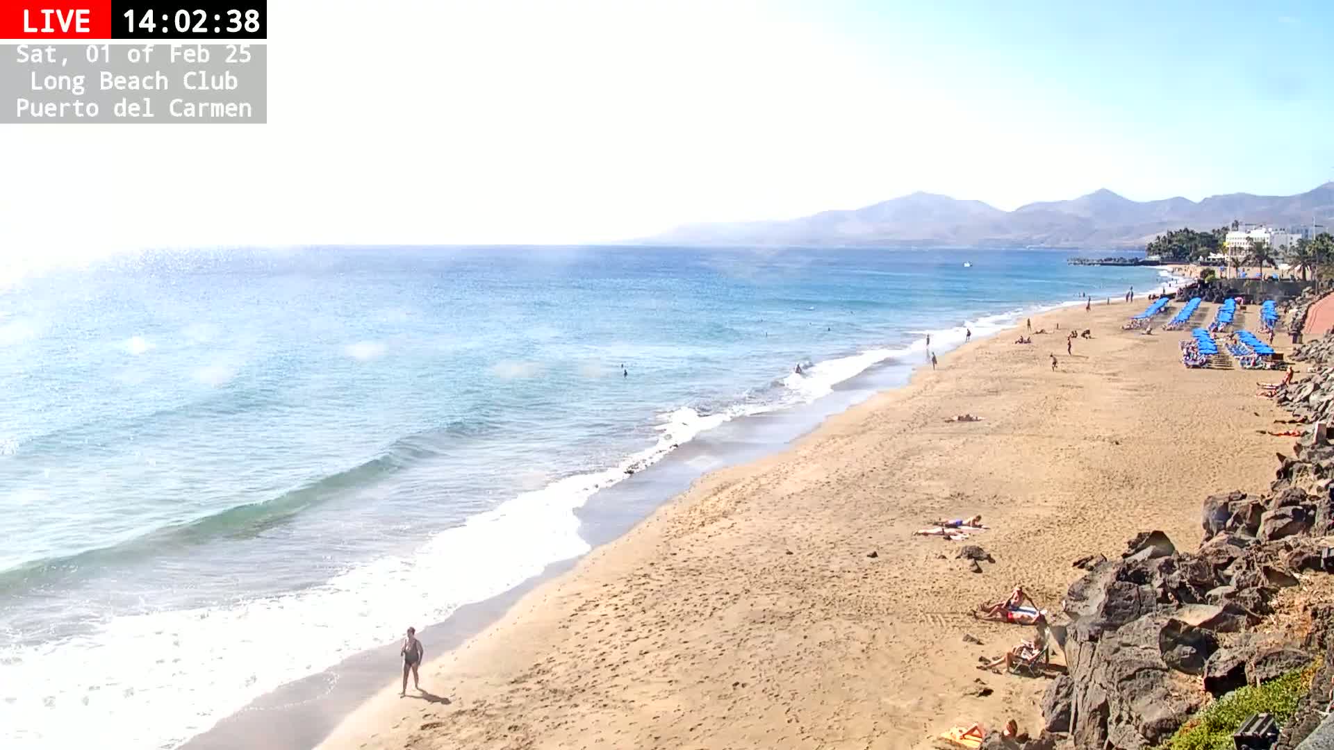 A sandy beach with a few people on it and rows of blue beach chairs is bordered by calm, light blue ocean waves under a sunny sky, with mountains in the distance.