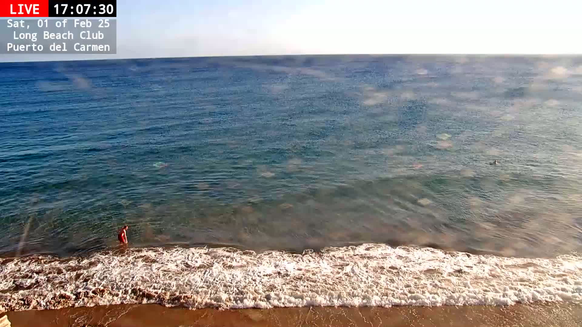 A person wades in relatively calm, clear ocean water near a sandy beach on a sunny day.