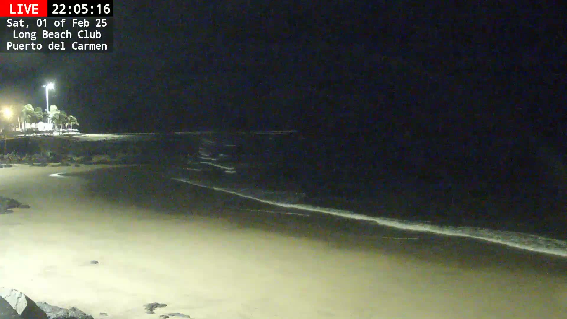 A nighttime view of a sandy beach with gentle waves lapping the shore under a dark sky.