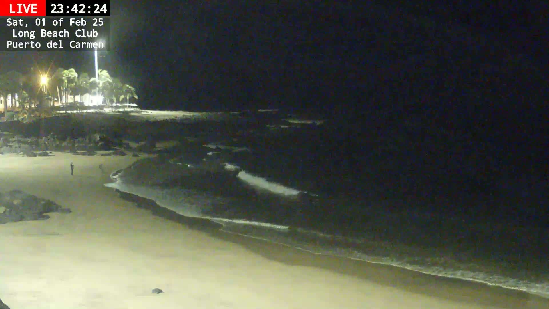 A dark sandy beach at night with gentle waves lapping the shore, and a single person visible in the distance.