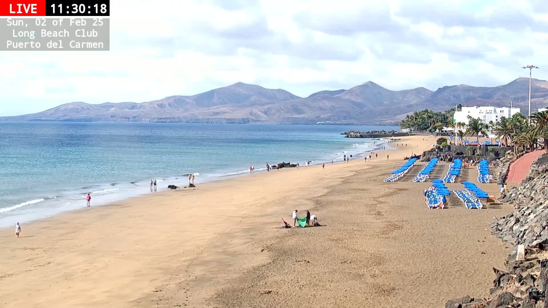 A sandy beach with blue umbrellas and lounge chairs stretches along a calm ocean, backed by mountains under a partly cloudy sky.