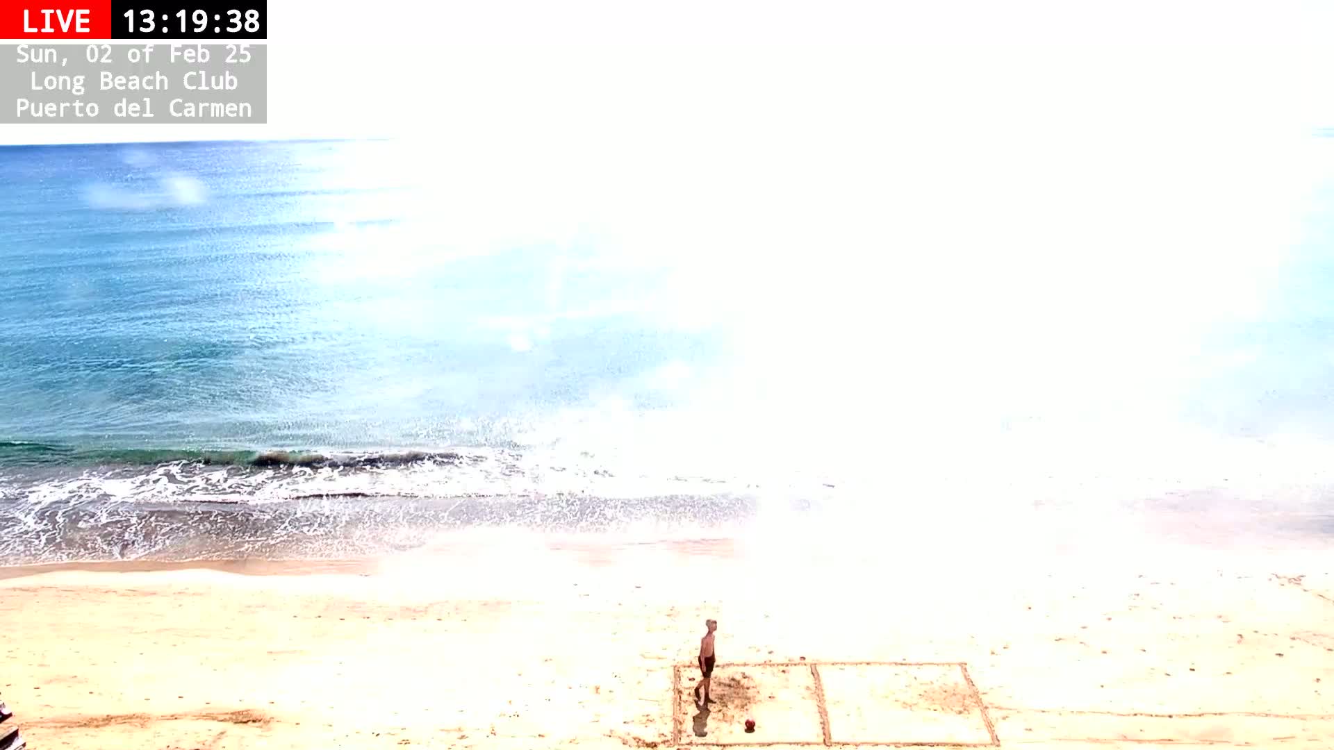 A man stands on a sandy beach next to a square drawn in the sand, overlooking a calm, light-blue ocean under a bright, mostly clear sky.