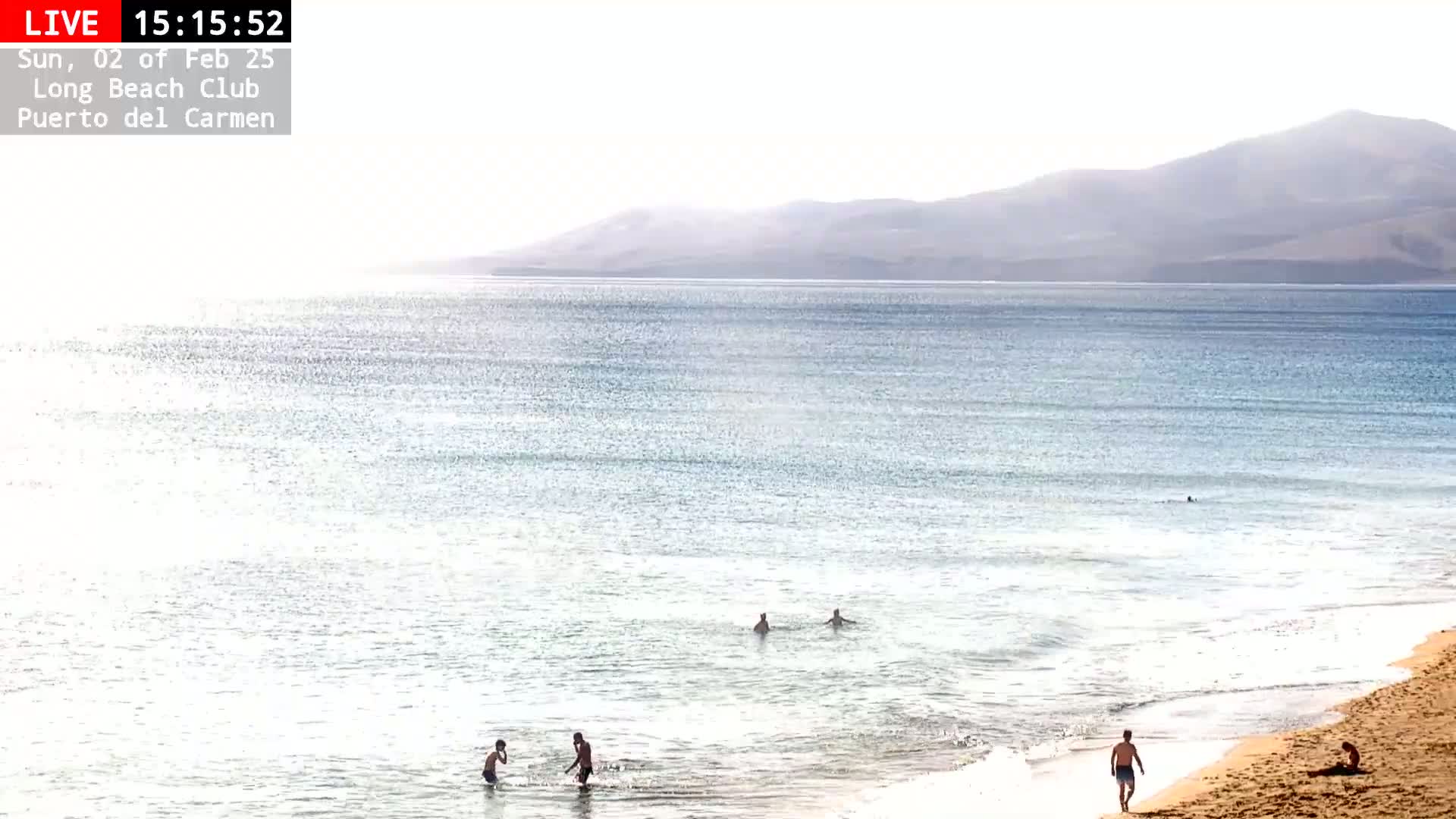 A sandy beach with several people swimming and walking near calm, light blue water under a bright, hazy sky, with mountains visible in the distance.