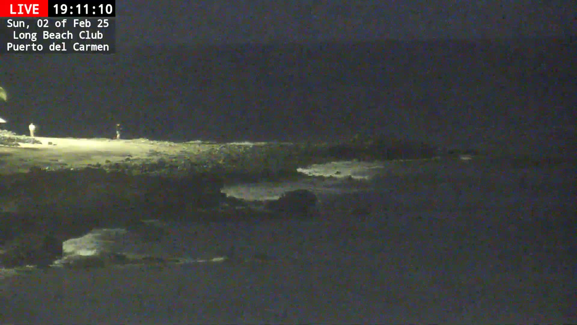 A dark, nighttime beach scene shows a rocky shoreline with gentle waves lapping against it, and two figures standing on the sand.