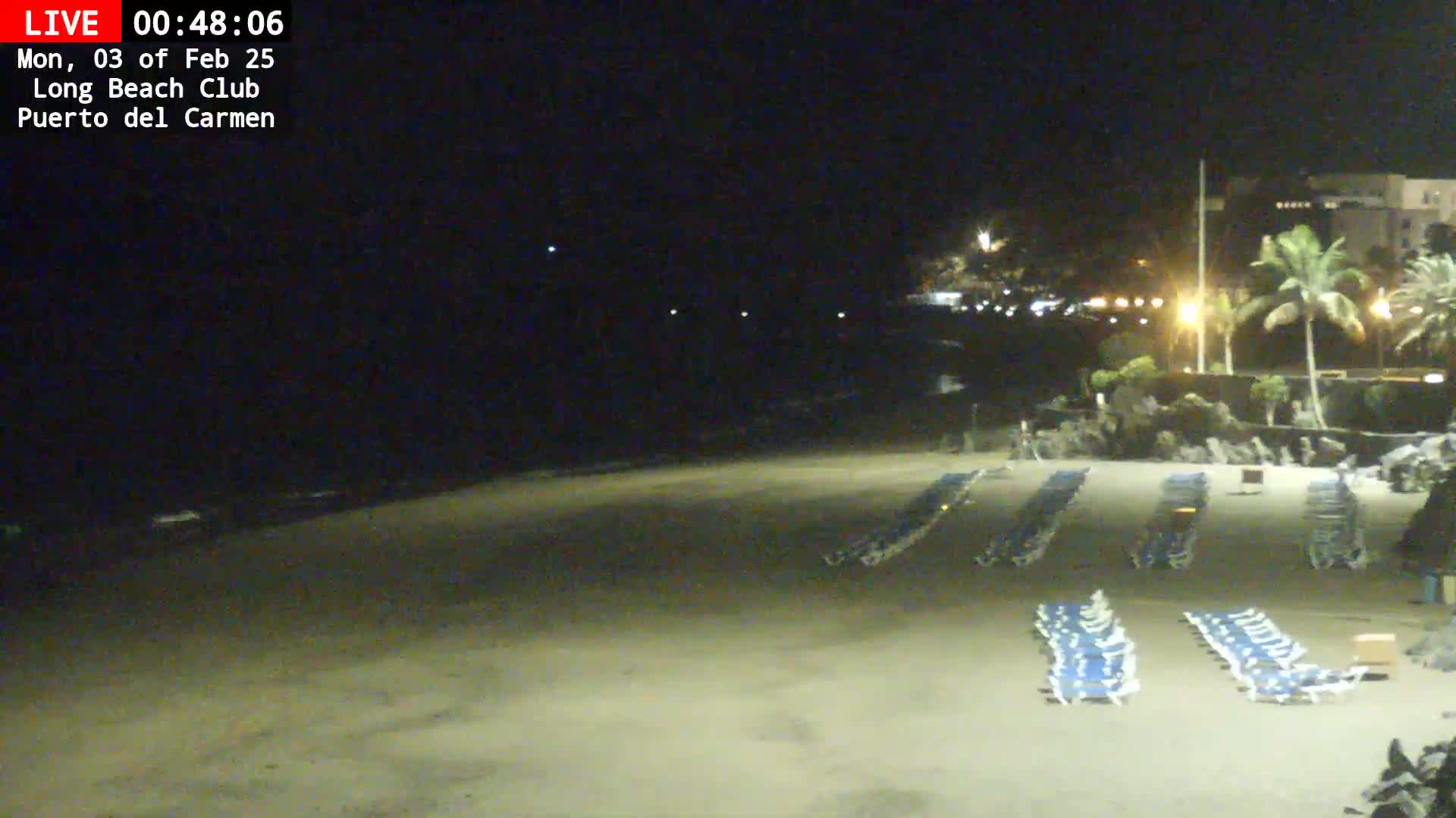 A dark sandy beach at night, lined with rows of empty lounge chairs, is illuminated by artificial lights under a clear sky.