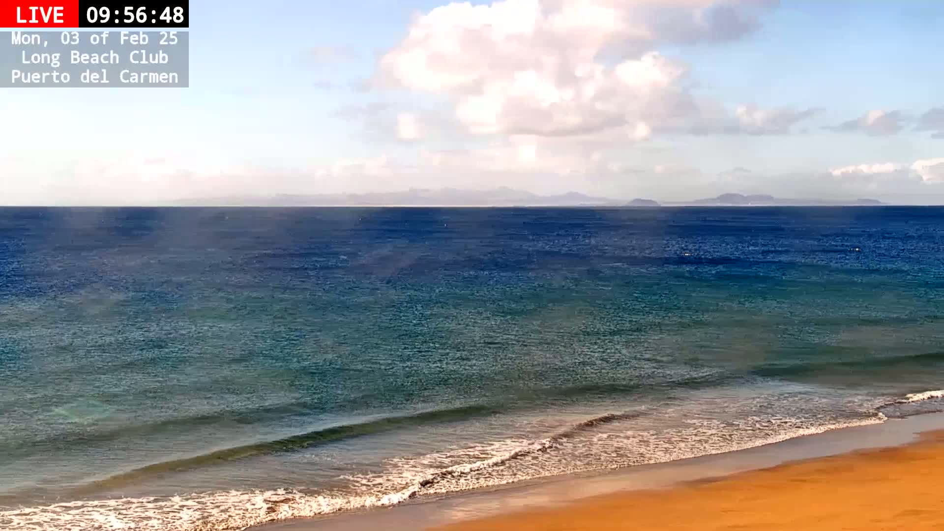A sandy beach meets a calm, blue ocean under a partly cloudy sky.