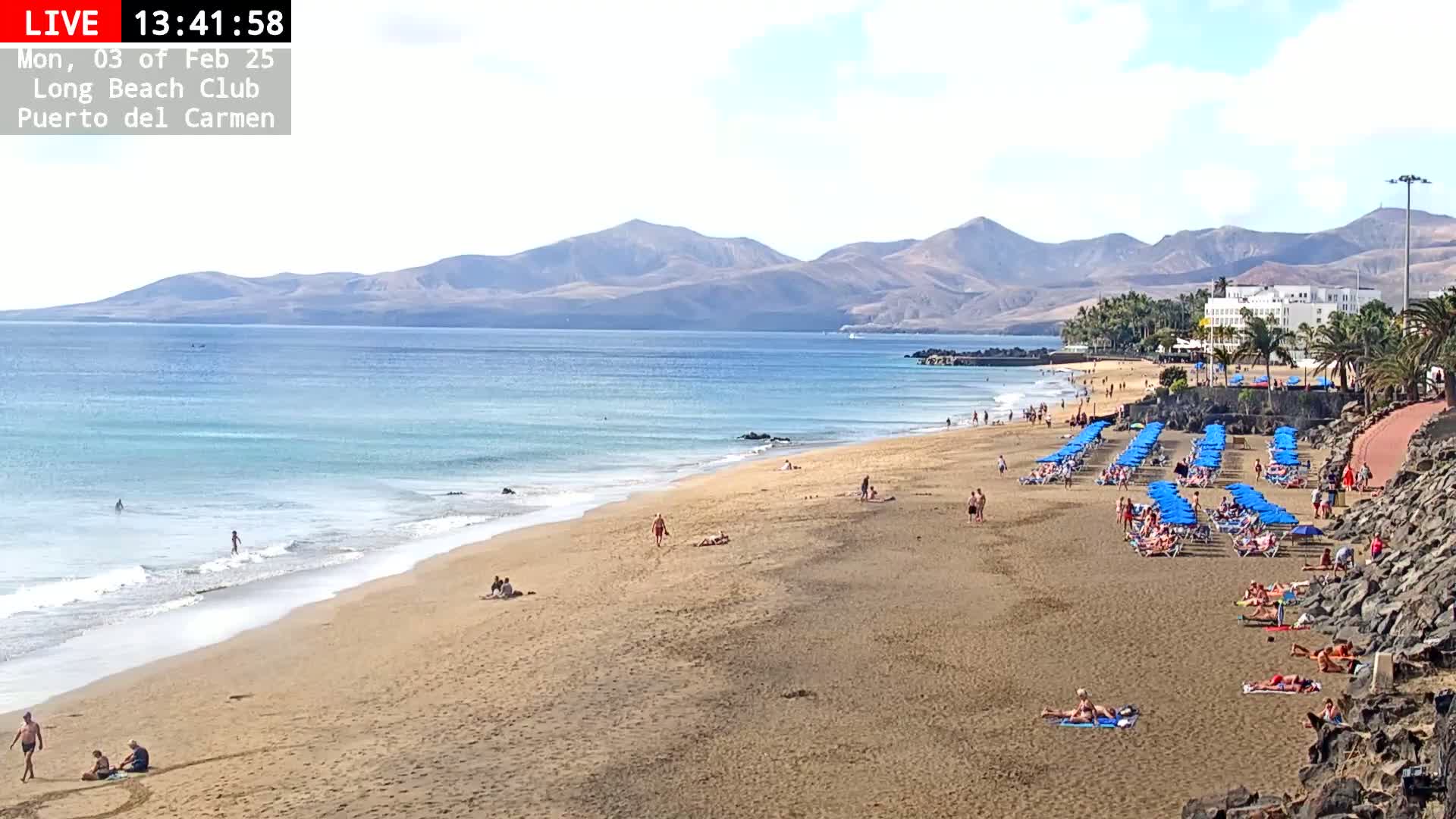 A sandy beach with blue umbrellas and scattered people stretches along a calm ocean, backed by mountains under a clear sky.