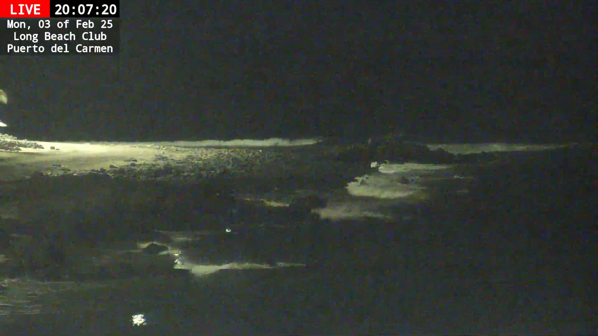 A dark, nighttime view of a rocky shoreline with waves gently lapping against the shore under a clear sky.