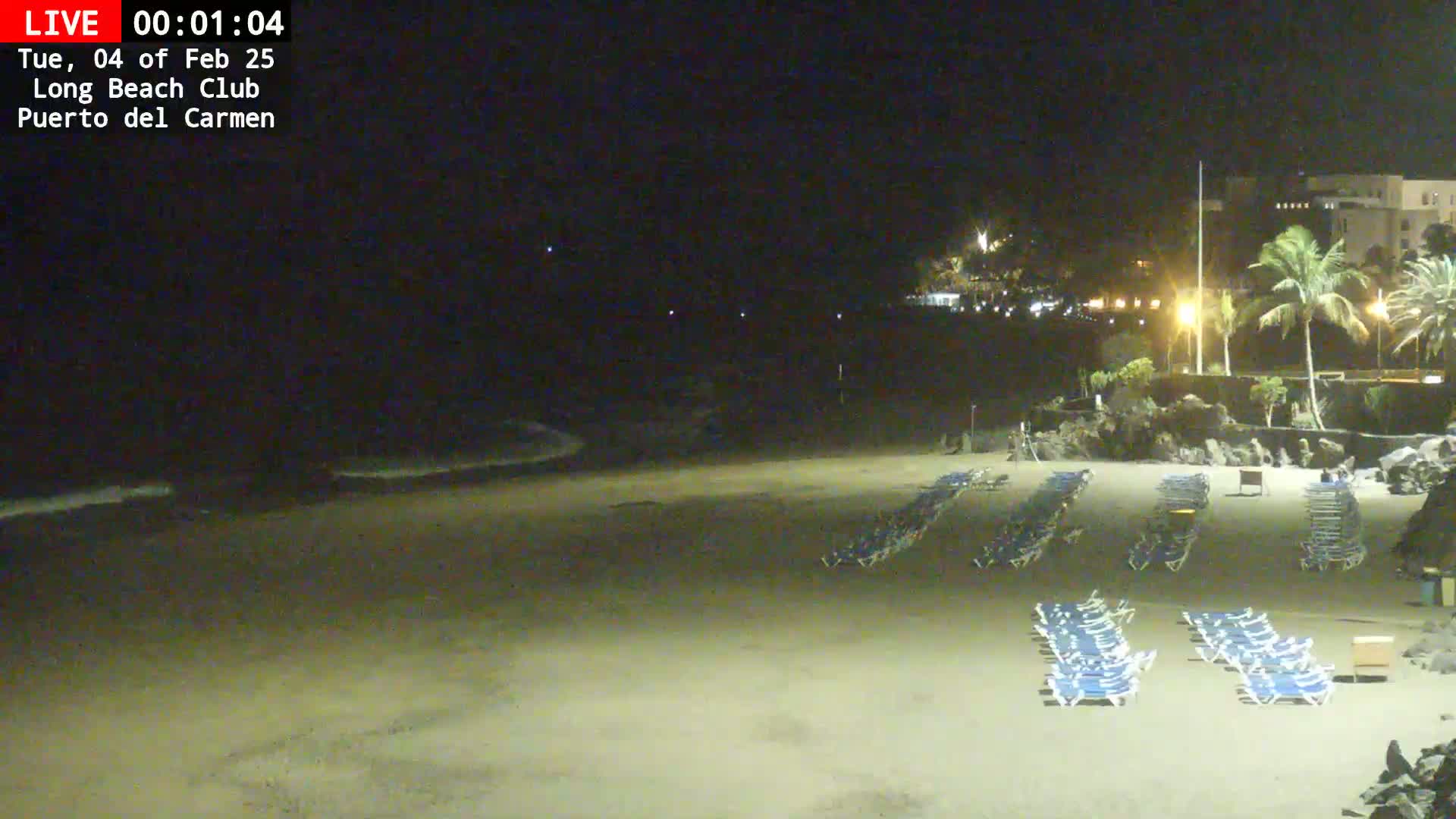 A dark sandy beach at night shows rows of empty lounge chairs and a calm ocean under a clear, dark sky.