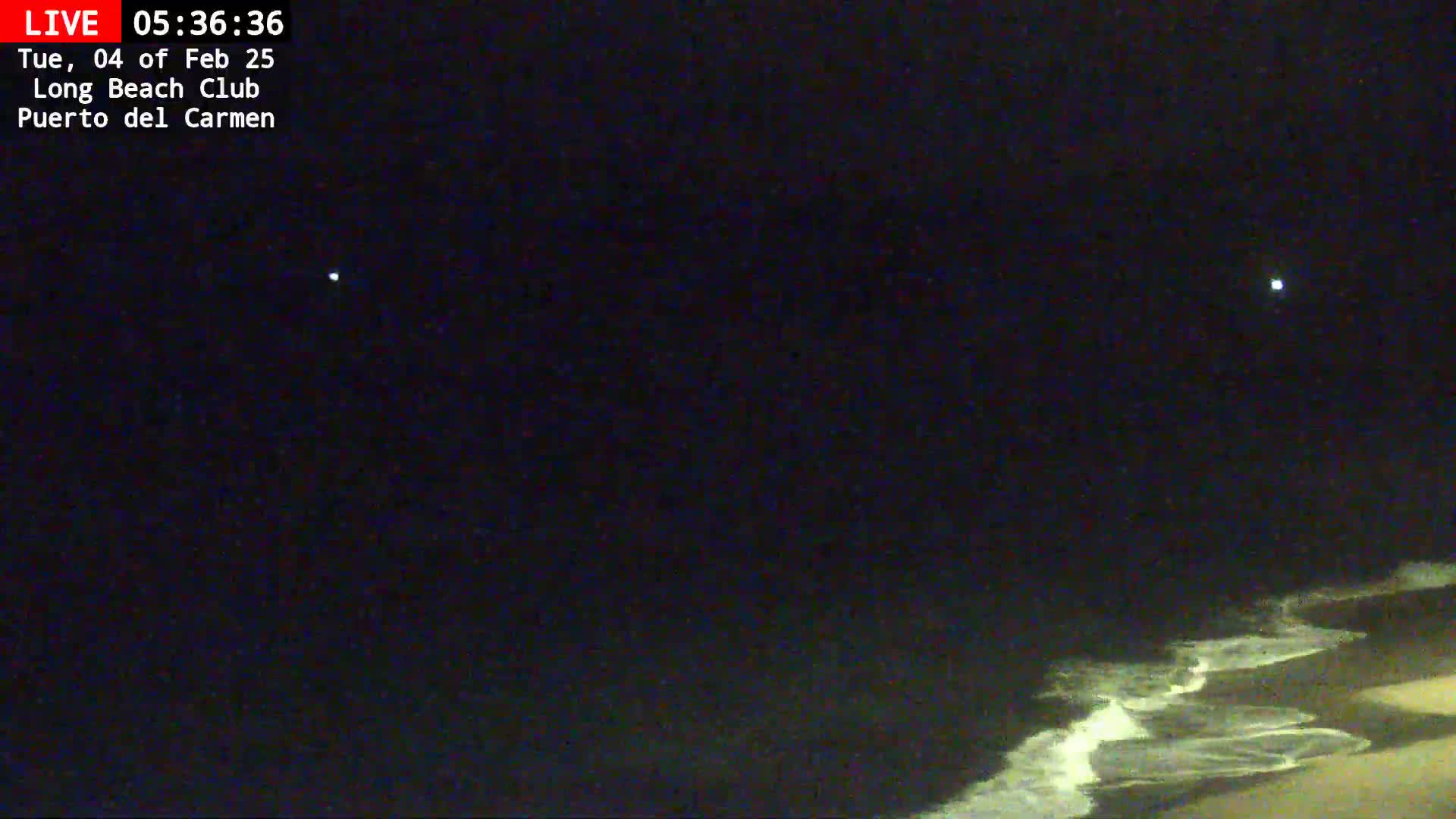 A dark night scene shows a beach with waves lapping the shore under a mostly clear sky with a few visible lights.