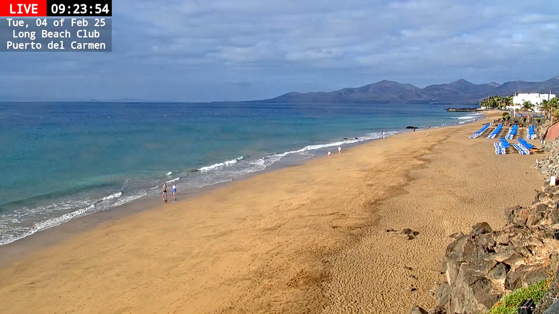 A sandy beach with gently lapping waves stretches towards a mountainous coastline under a partly cloudy sky.