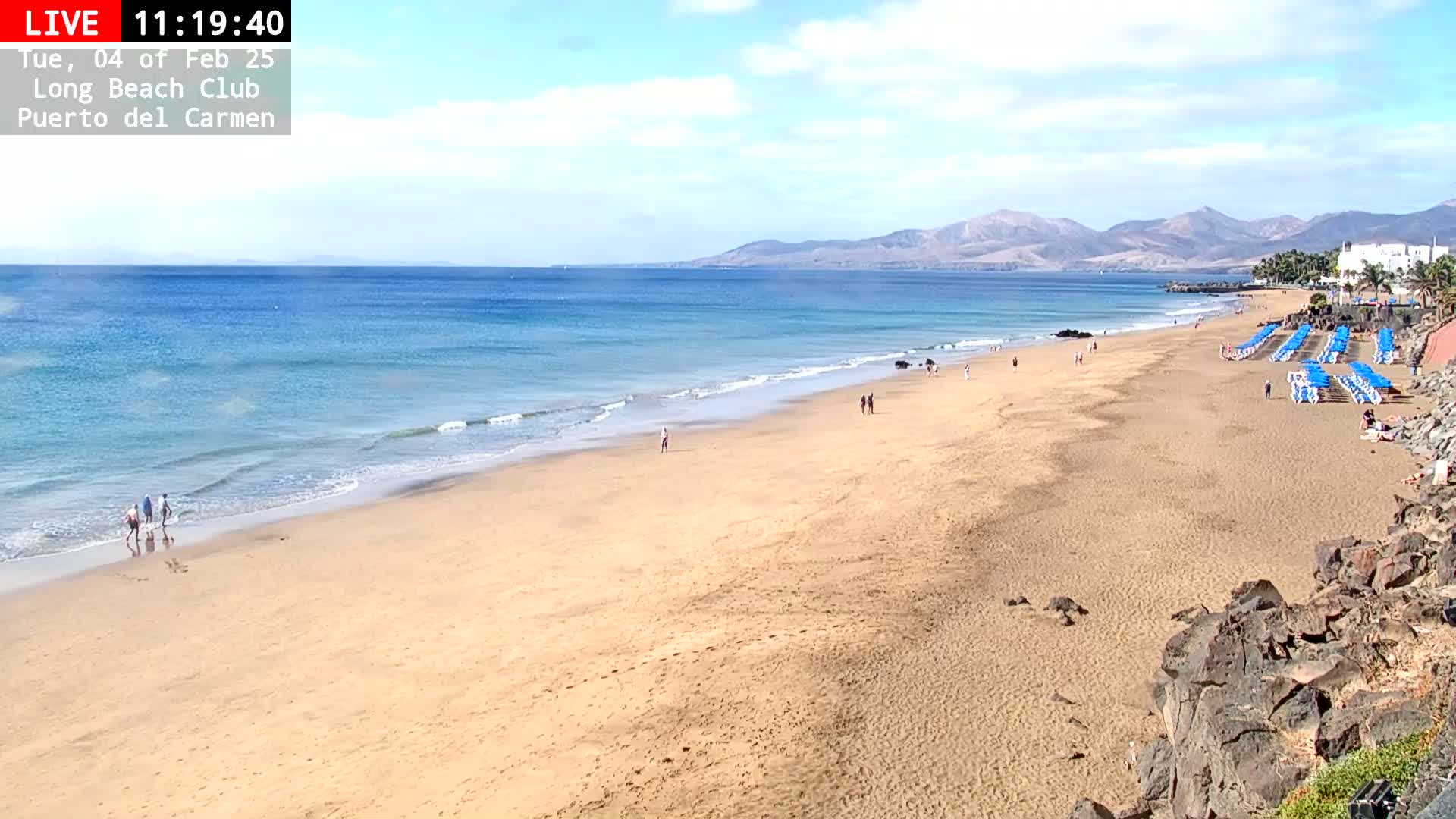 A long sandy beach stretches to a calm, blue ocean under a mostly sunny sky, with mountains visible in the distance and a few people scattered along the shore.