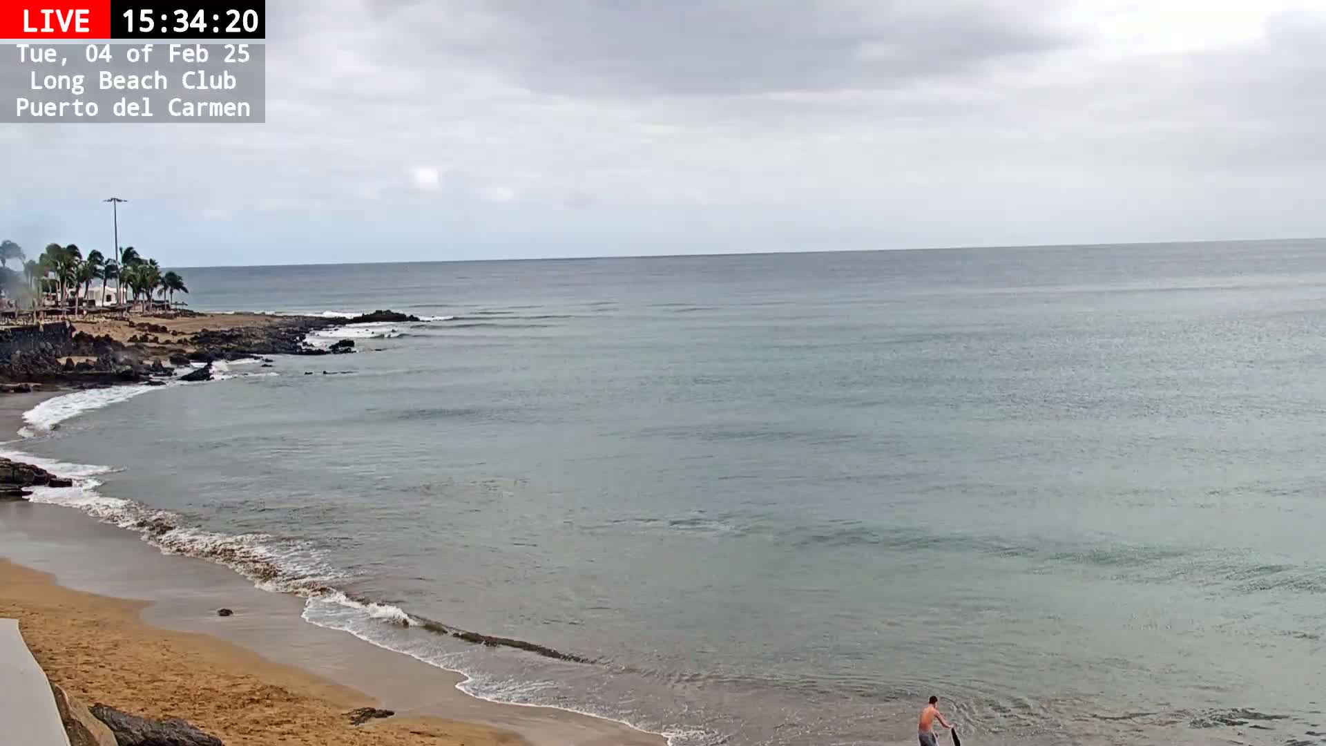 A person stands in the shallow water of a sandy beach on an overcast day, with a calm ocean extending to the horizon.